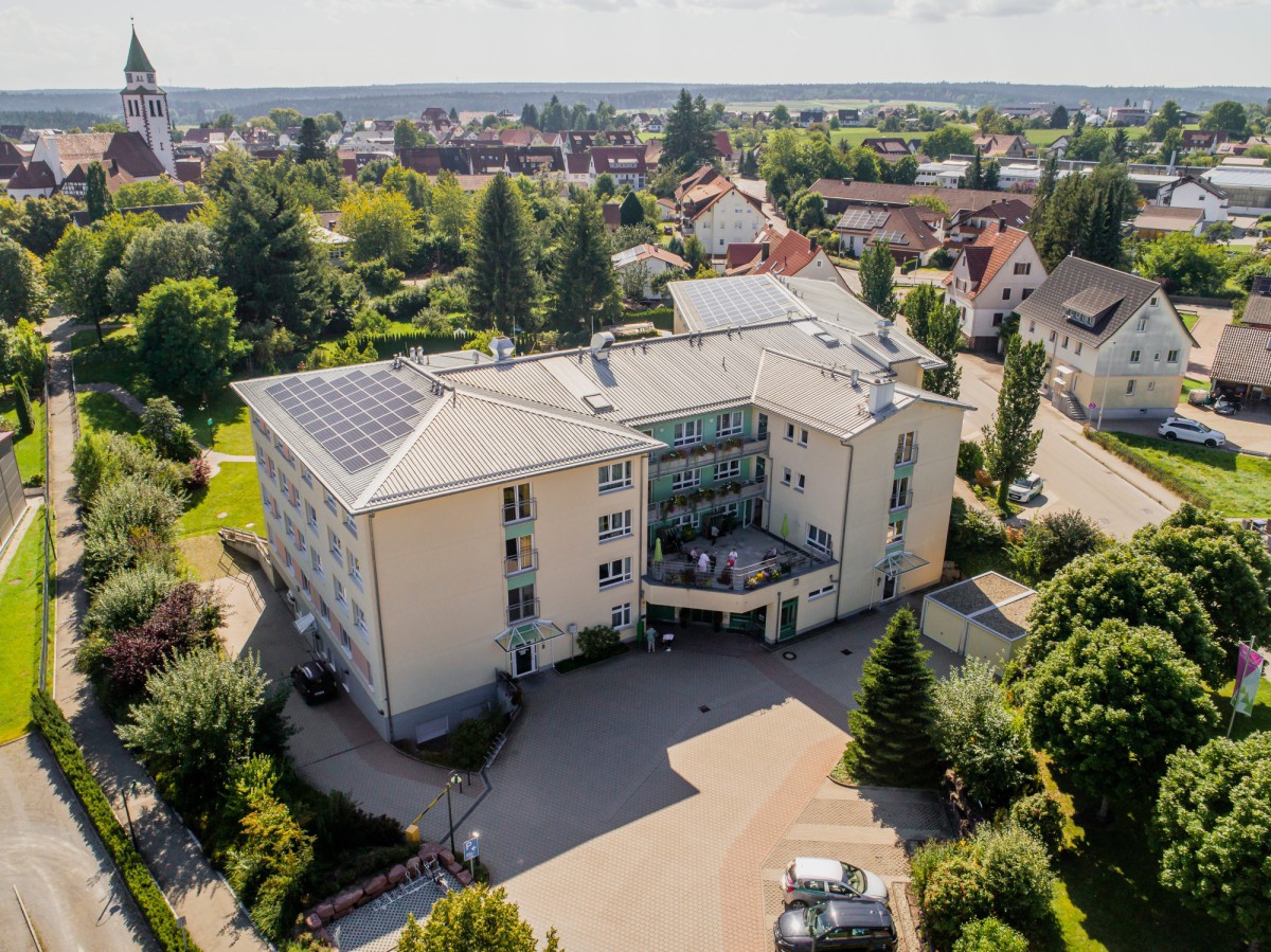 Luftaufnahme des Seniorenstifts AM KURGARTEN mit Solaranlage auf dem Dach mit Kirche und Häusern im Hintergrund. Das moderne Gebäude hat Balkone und eine gepflegte Umgebung mit Bäumen und Parkplätzen.