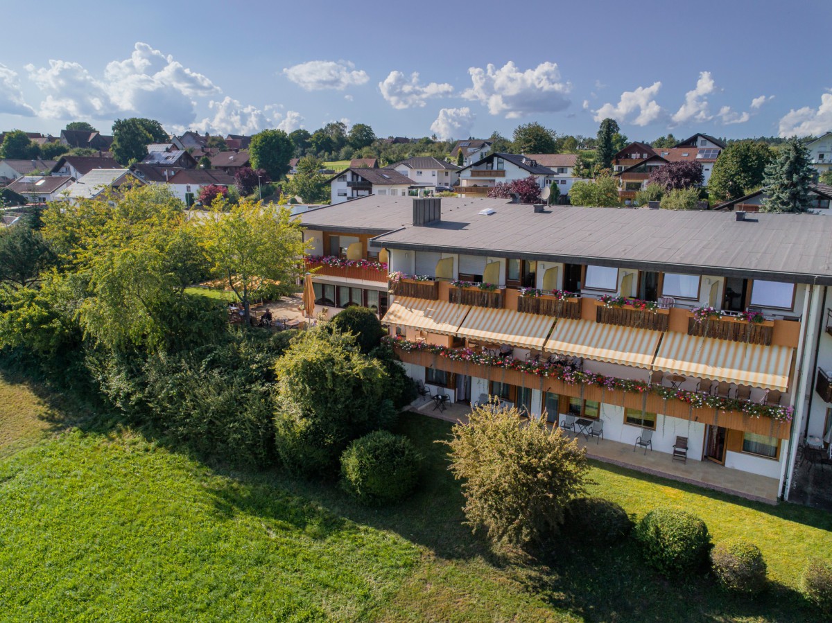 Luftaufnahme des Seniorenstifts MANDELBERG mit Holzbalkon und roten Blumenkästen, umgeben von grünem Rasen, Büschen und Bäumen, mit Häusern im Hintergrund unter blauem Sommerhimmel.