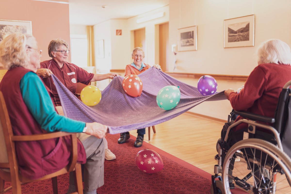 Mehrere ältere Menschen sitzen im Gemeinschaftsraum und spielen gemeinsam mit einem gespannten Tuch und bunten Luftballons.