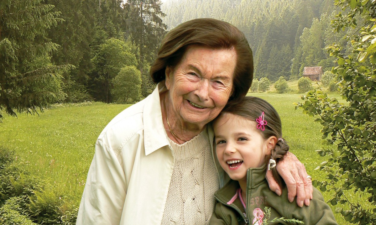 Ältere Frau umarmt lächelndes junges Mädchen mit Zöpfen vor einer grünen Wiese und Waldkulisse. Beide strahlen vor Freude und Verbundenheit.