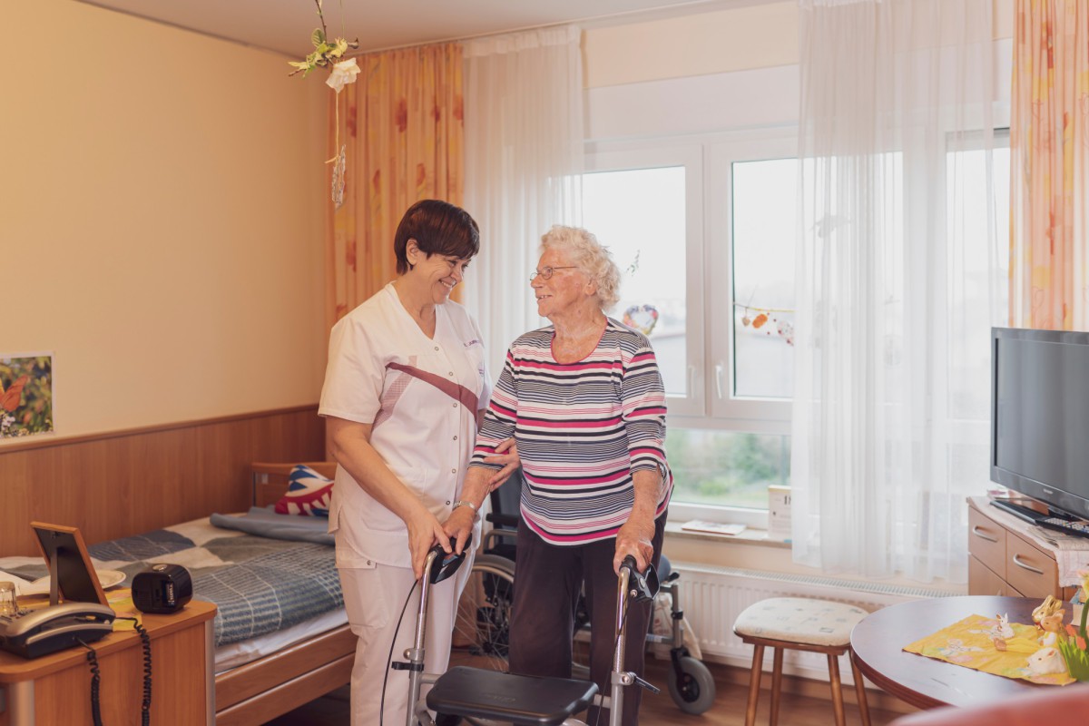 Eine Pflegekraft unterstützt eine ältere Dame mit einem Rollator in einem hellen, gemütlichen Zimmer mit Holzvertäfelung und Vorhängen.