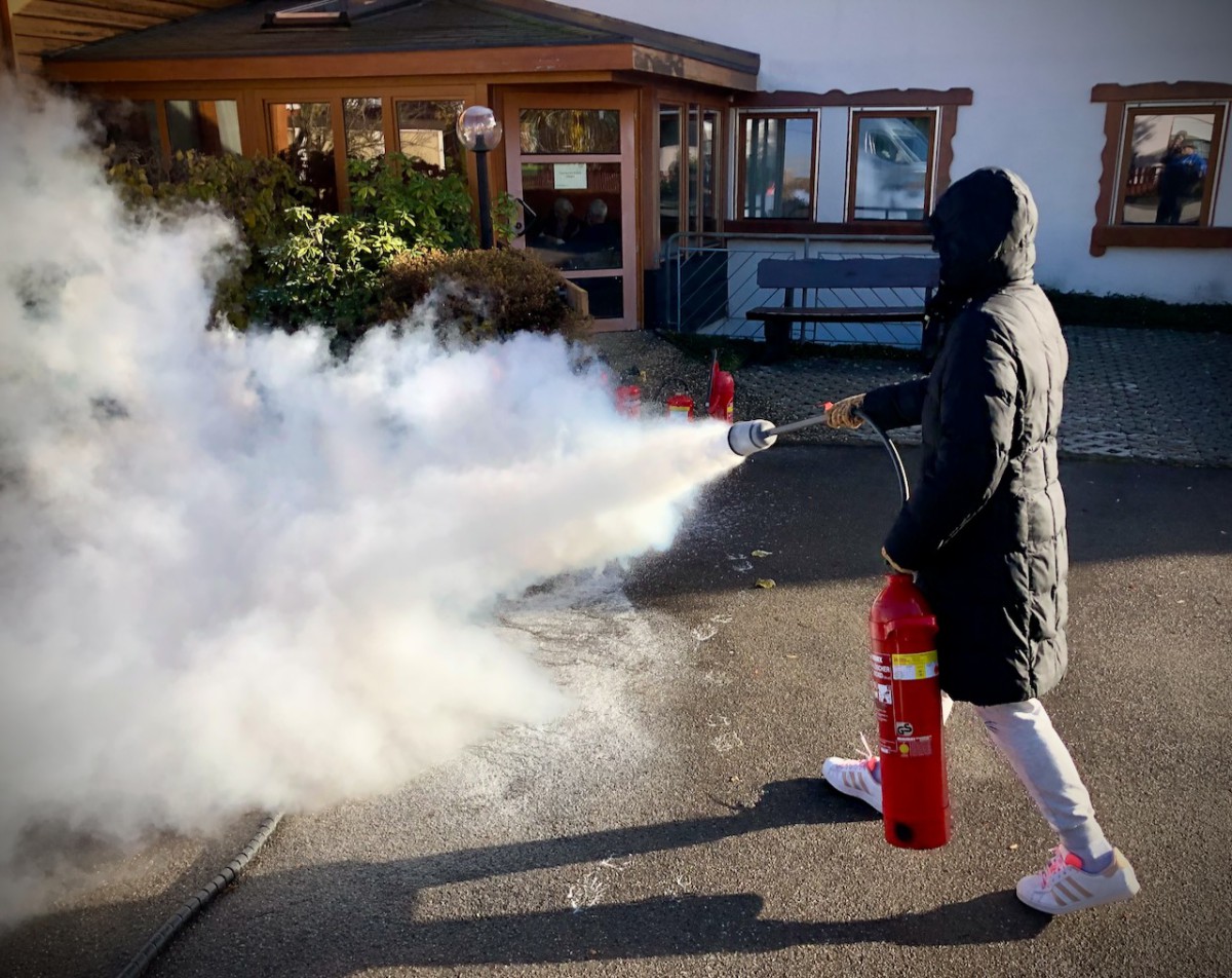 Eine Person in dunkler Winterjacke und Kapuze bedient einen roten Feuerlöscher, der eine große weiße Rauchfahne auslöst, vor einem Gebäude mit Holzüberdachung und Glasfenstern.