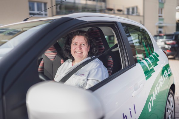 A smiling person sitting in a white car with KSD logo, buckled up and ready to drive.