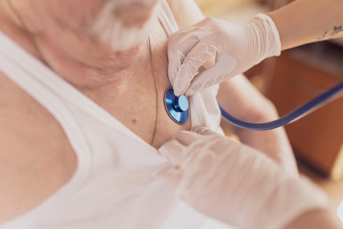 Arzthand mit Handschuhen untersucht einen Patienten mit einem blauen Stethoskop, während der Patient ein weißes Oberteil trägt.