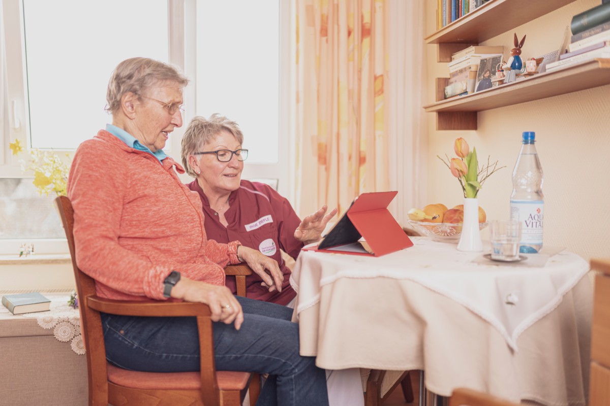 Zwei Personen sitzen an einem Tisch mit einem roten Tablet, umgeben von gemütlicher Wohnzimmeratmosphäre mit Büchern, Vorhängen und Blumen.