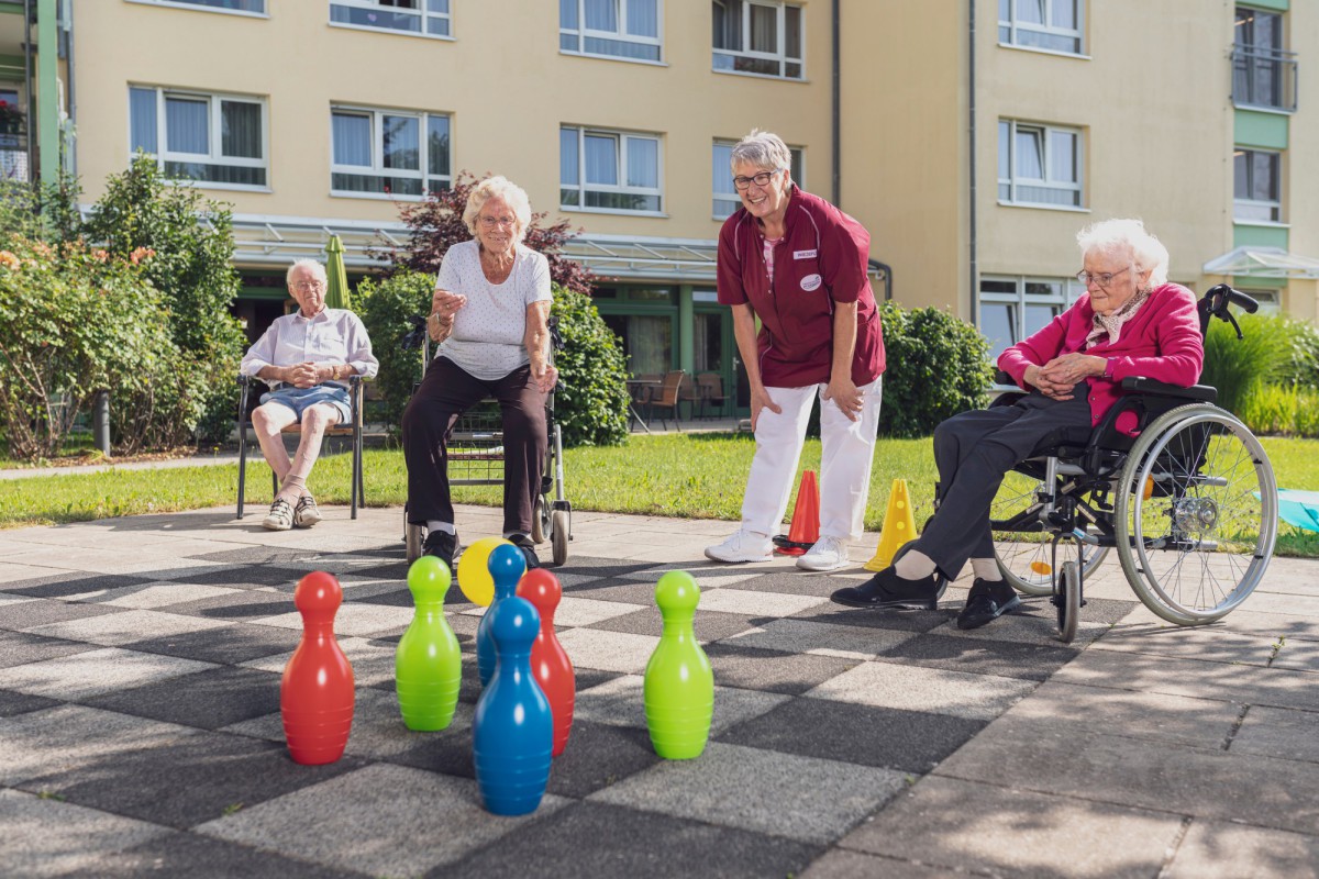 Drei Seniorinnen und Senioren spielen Bowling auf einem Schachbrettmuster im Freien, ein Mitarbeiterin in Rot-Weiß assistiert dabei.