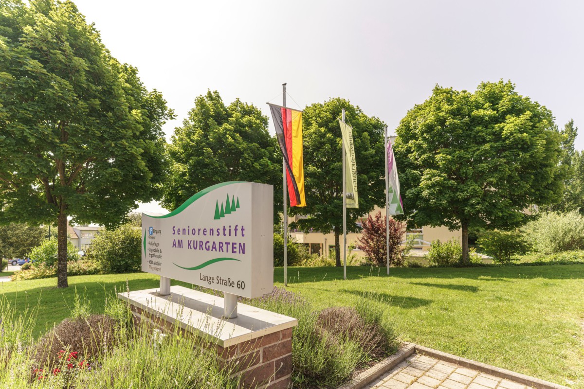 Sign of the senior residence Am Kurkarten with German flag in front of green garden with trees and flower beds.