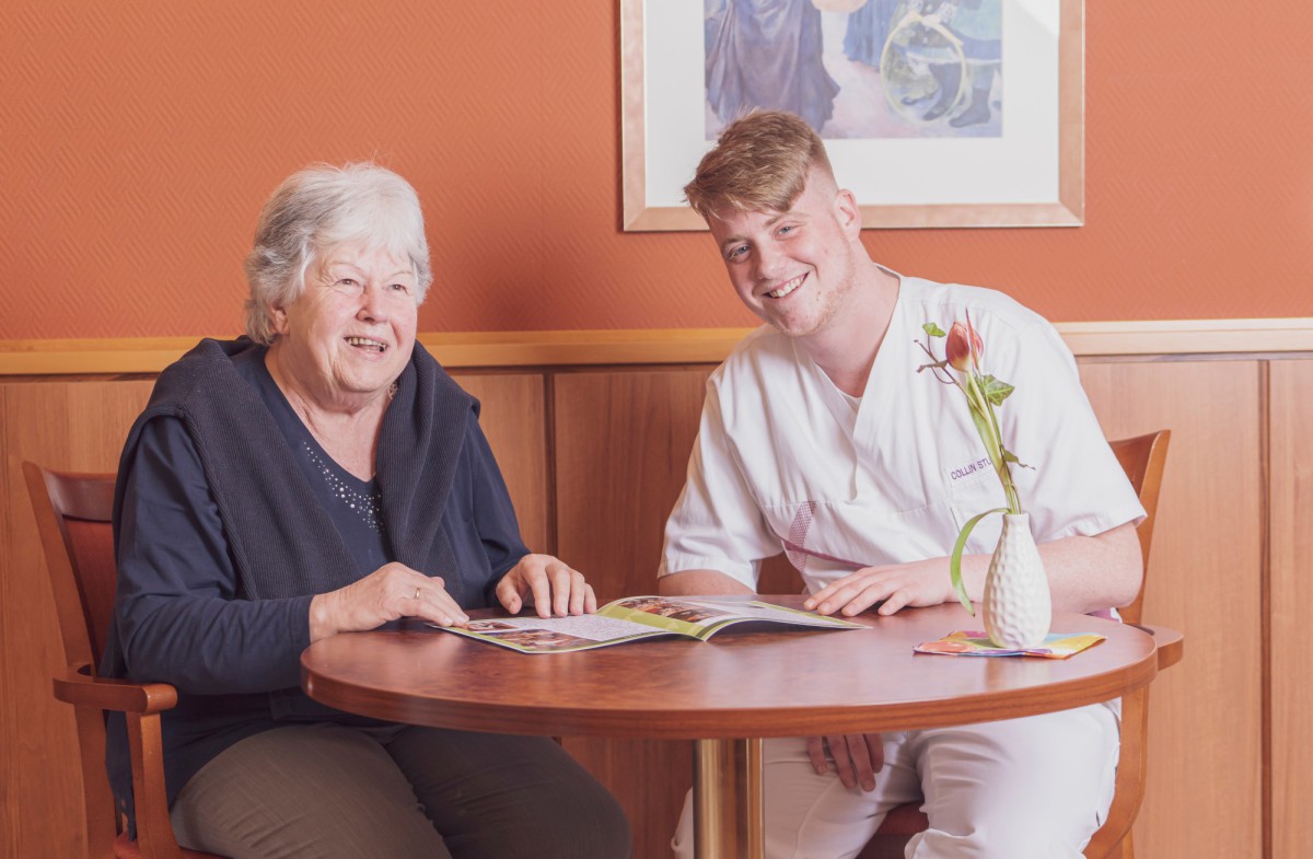 Older woman in dark blue blouse and younger man in white shirt smiling at a round wooden table, with orange-toned wall and framed artwork in background.