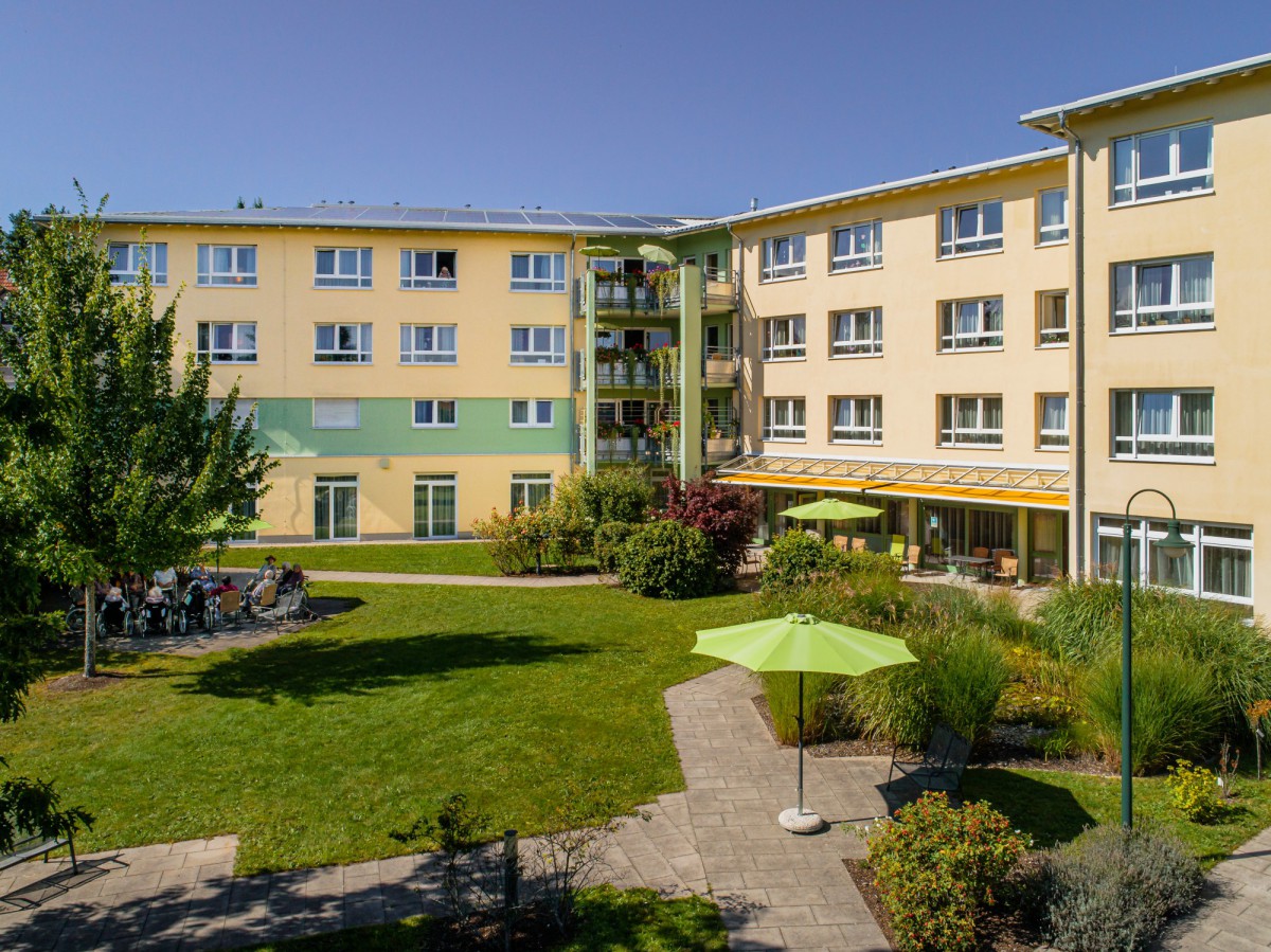 Aerial view of a senior residence at AM KURGARTEN with solar panels on the roof, modern building with balconies and well-maintained surroundings with trees and parking spaces.