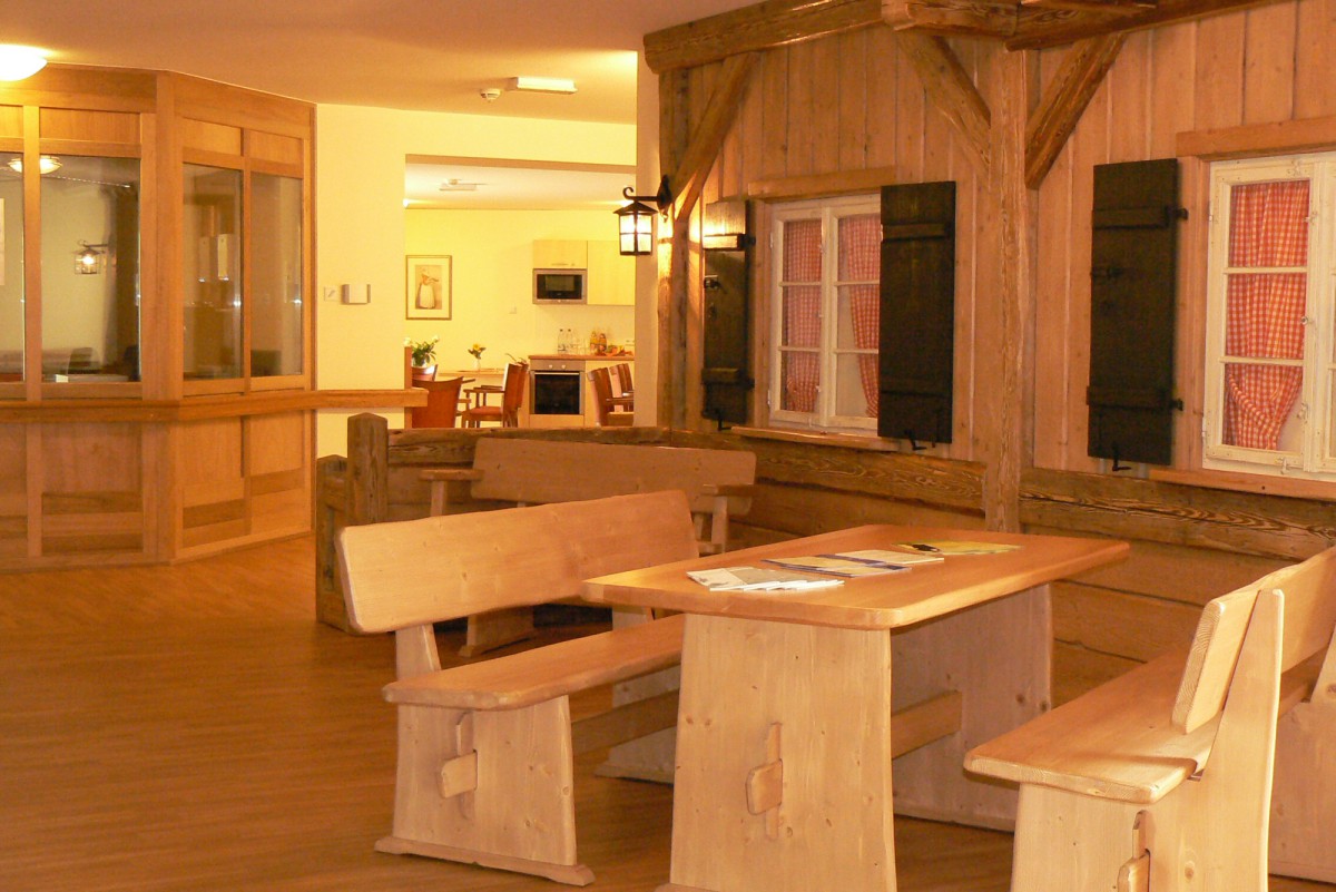 Rustic interior with wooden furniture, featuring a wooden bench and table by a window with black shutters, warm and bright atmosphere with wood paneling, connecting to an adjacent room.