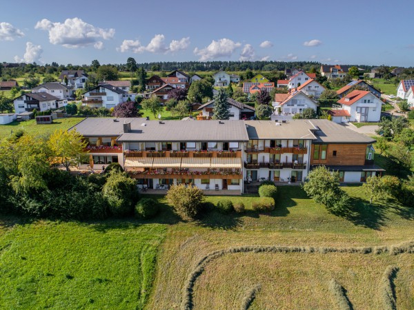 Aerial view of Mandelberg senior residence with balconies, surrounded by green trees and meadows, with a residential area in the background and blue sky.