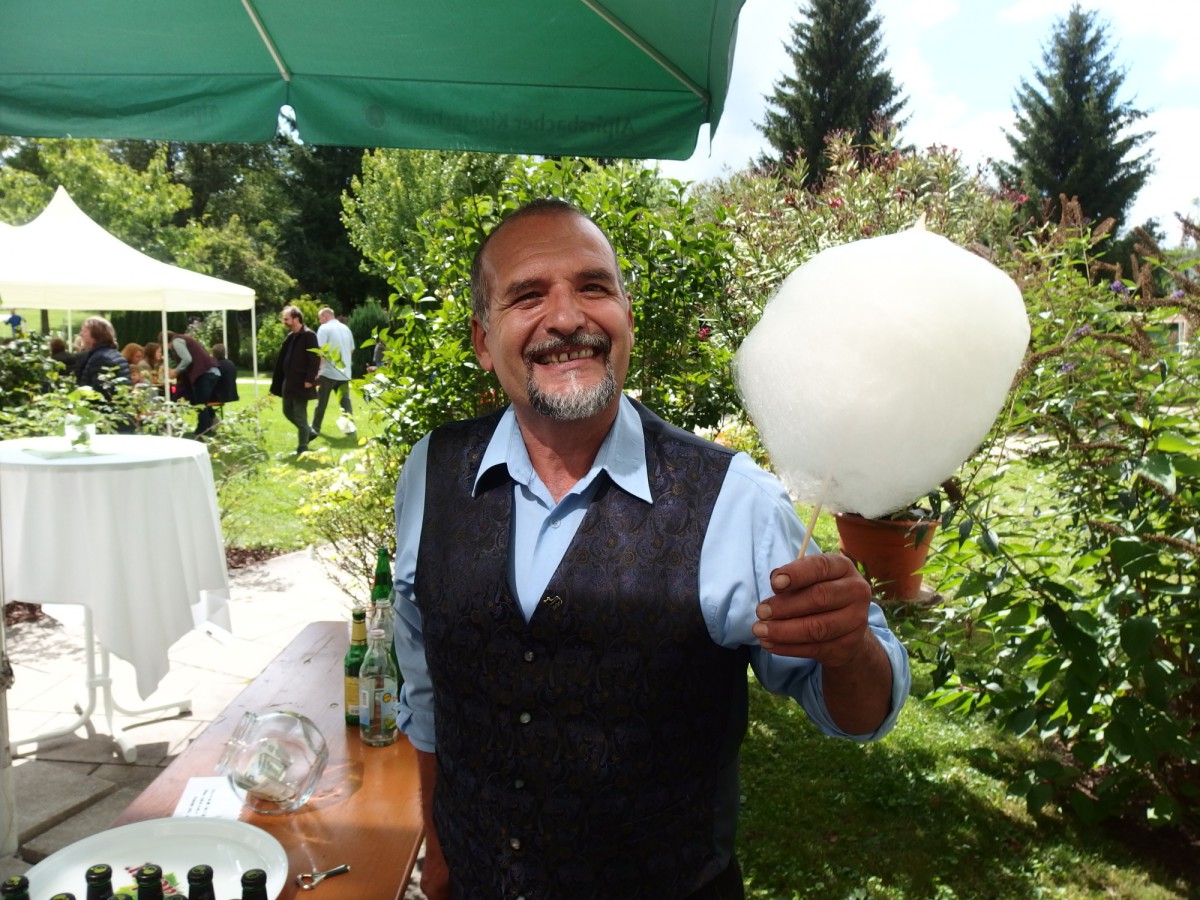 Ein freundlicher Mann in Weste und Hemd lächelt breit und hält Zuckerwatte in die Kamera.