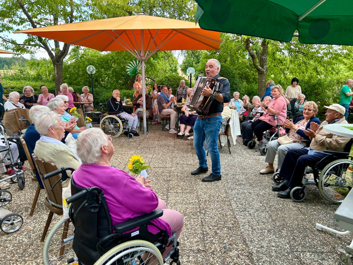 Seniorinnen und Senioren sitzen unter orangefarbenen und grünen Sonnenschirmen, während ein Mann Akkordeon spielt und eine entspannte Atmosphäre im Freien schafft.