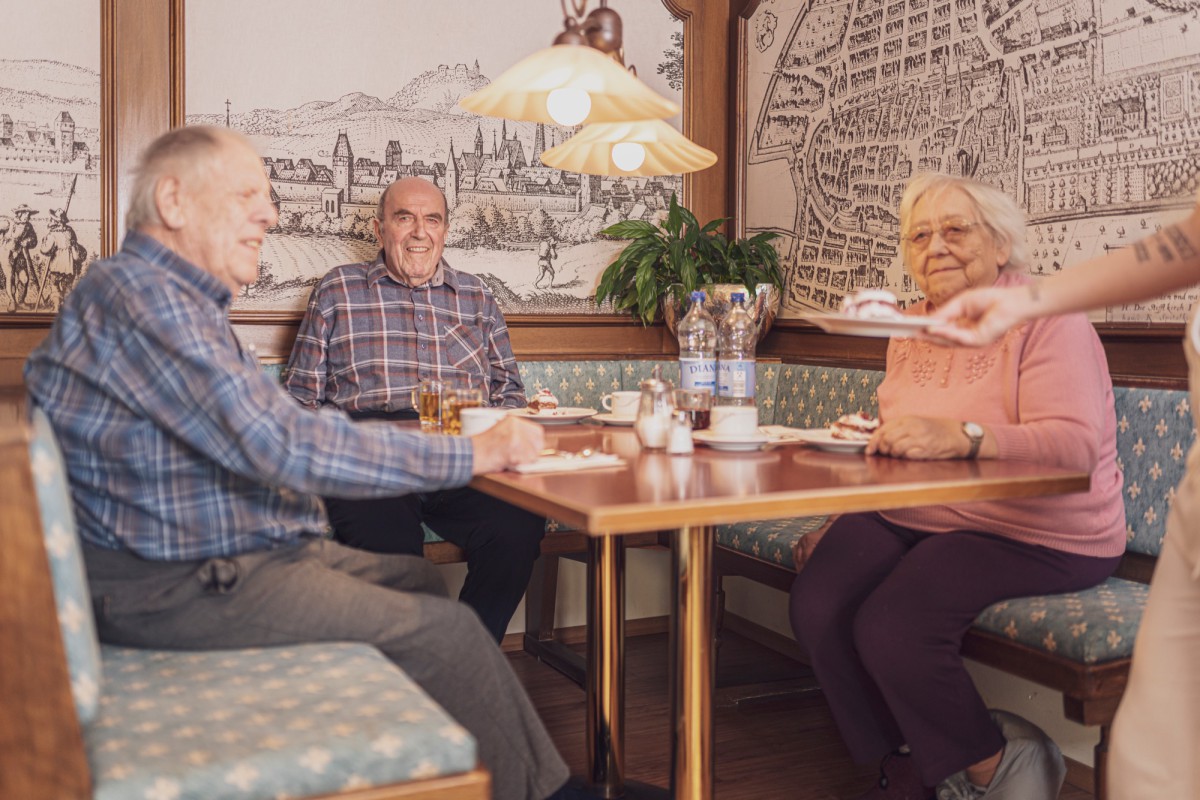 Drei ältere Menschen sitzen an einem Tisch in einem gemütlichen Café mit historischer Wanddekoration.