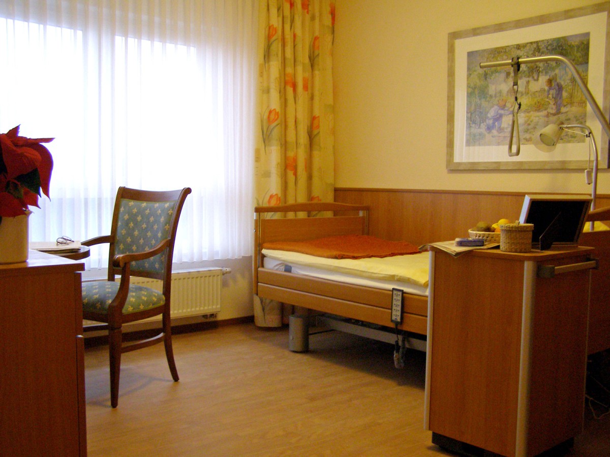 Zimmer mit einem Einzelbett, verstellbarem Nachttisch, Stuhl und einem Fenster mit Vorhängen. Ein Kunstdruck hängt an der Wand.