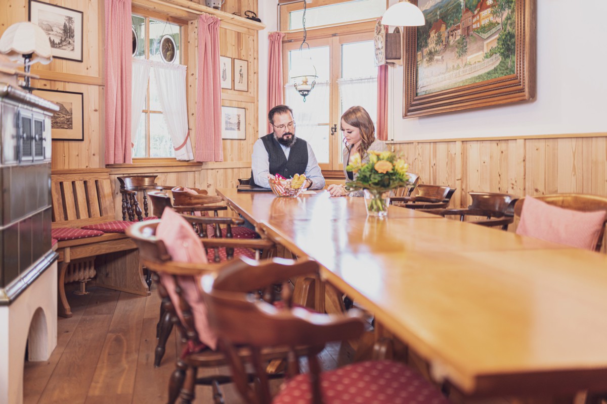Ein eleganter Gastraum mit Holzvertäfelung, in dem zwei Personen an einem langen Holztisch sitzen, umgeben von rustikalen Holzstühlen und Gemälden an den Wänden.