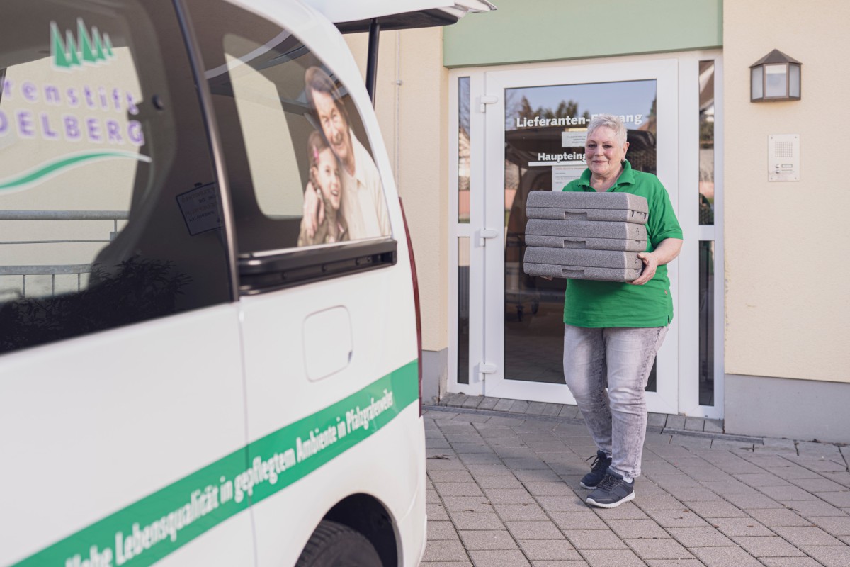 Eine Person in grünem T-Shirt trägt mehrere gestapelte Pakete vor einem weißen Lieferwagen mit grüner Beschriftung. Die Szene spielt sich vor einem Gebäudeeingang mit Glastür ab.