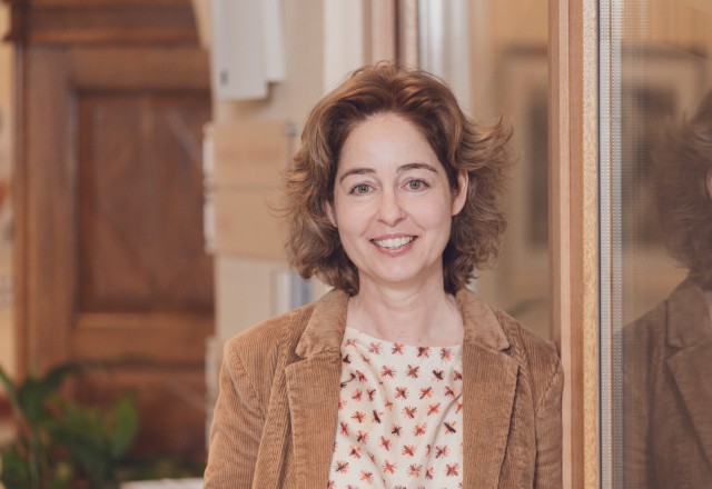 Lächelnde Frau mit gewellten braunen Haaren, trägt ein cremefarbenes Top mit Blumenmuster und eine braune Cordweste.