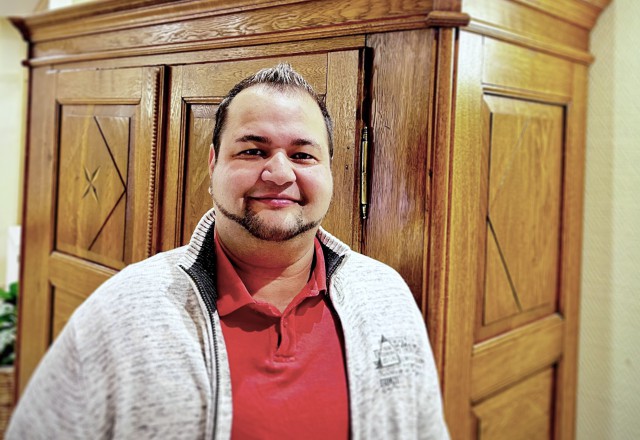 Mann in rotem Poloshirt und grau melierter Strickjacke lächelt freundlich in die Kamera, im Hintergrund eine großer Holzschrank.